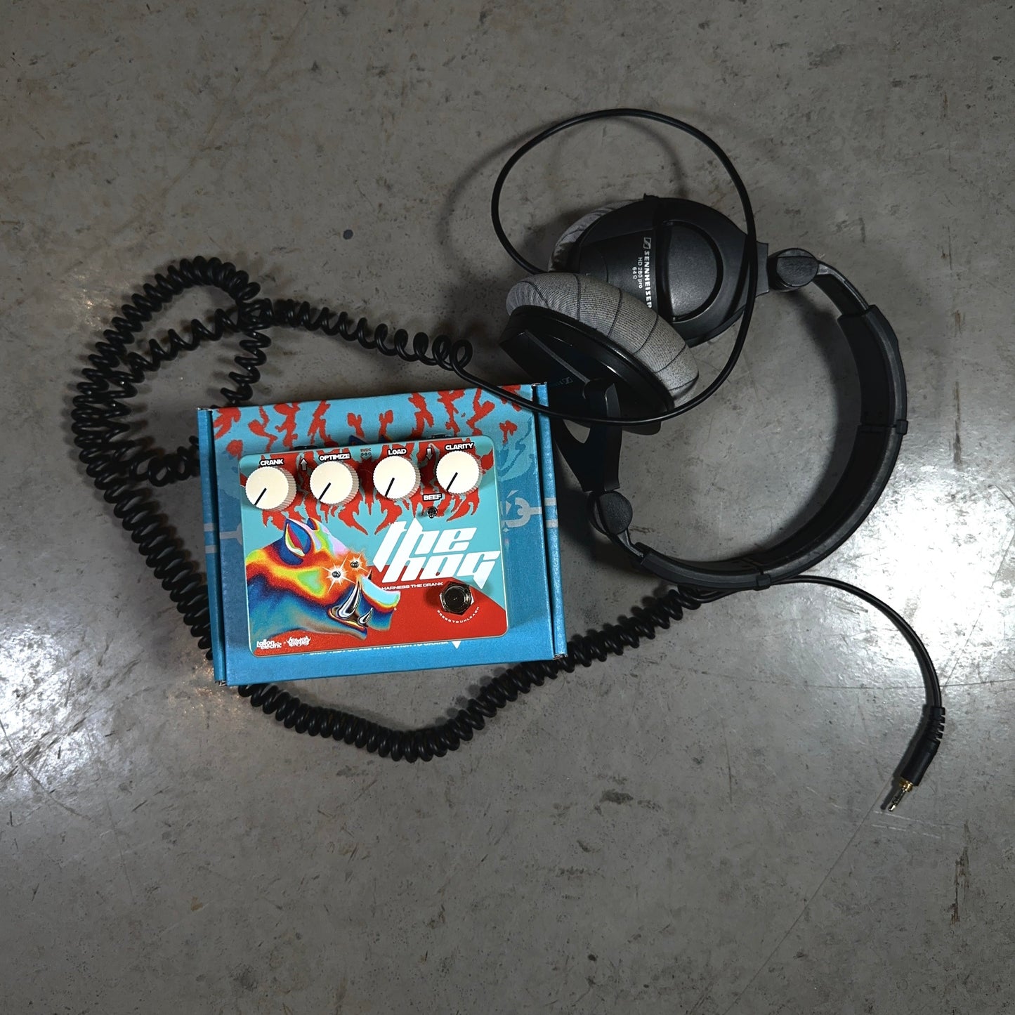 A pair of studio headphones and their cord tangle around The Hog guitar pedal by Tallon Electric and Bilmuri.